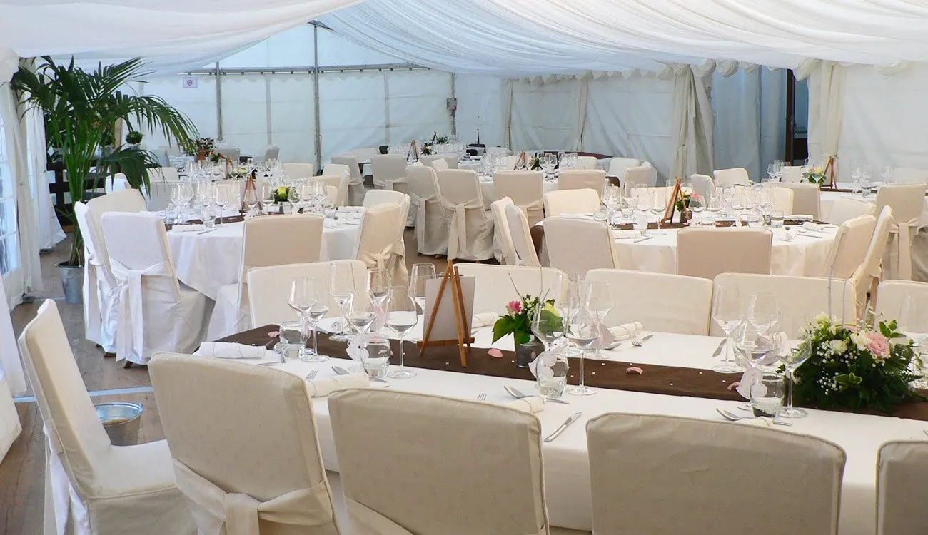 Tables rondes dressées, chaises housées de blanc, sous une tente